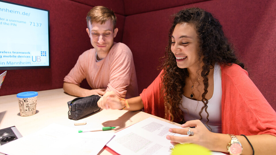 Zwei Studierende sitzen lachend an einem Tisch. Vor ihnen ausgedruckte Texte, Stifte und ein Kaffeebecher.